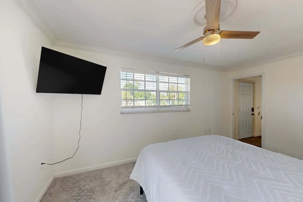 A bright bedroom with a bed, a wall-mounted television, and a window.