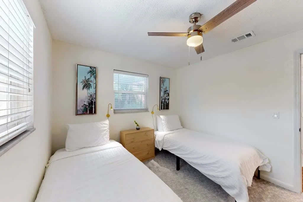A bright bedroom with two beds, a wooden nightstand, and a ceiling fan.