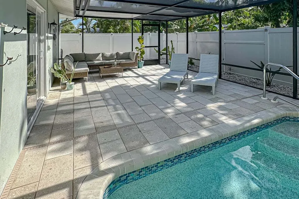 A paved patio beside a swimming pool has lounge seating beneath a covered frame.