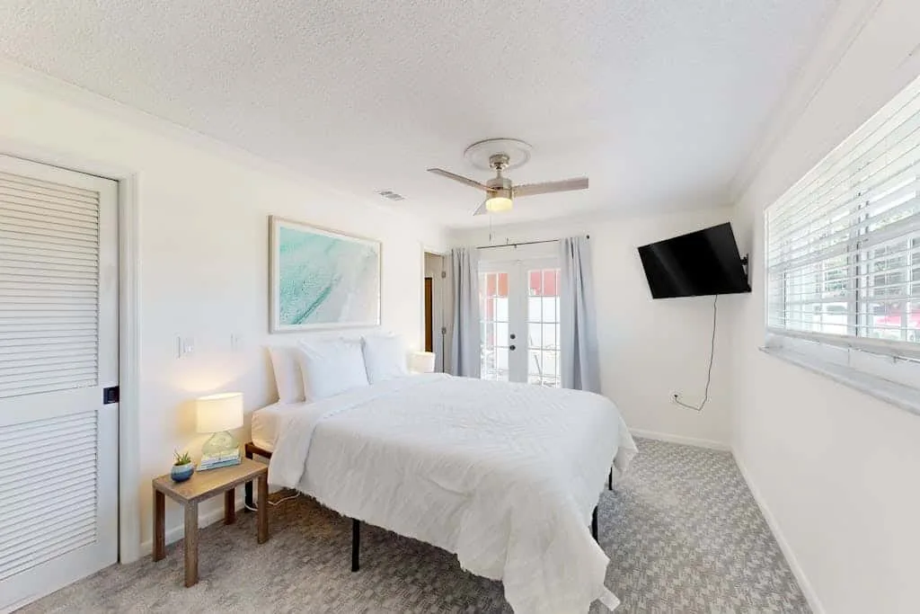 A bright bedroom with a white bed, bedside table, and wall-mounted television.