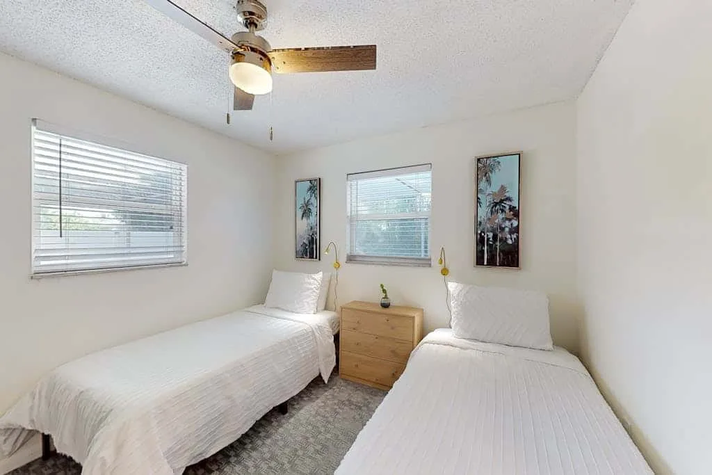 A bright bedroom with two beds, windows, and a small wooden nightstand.