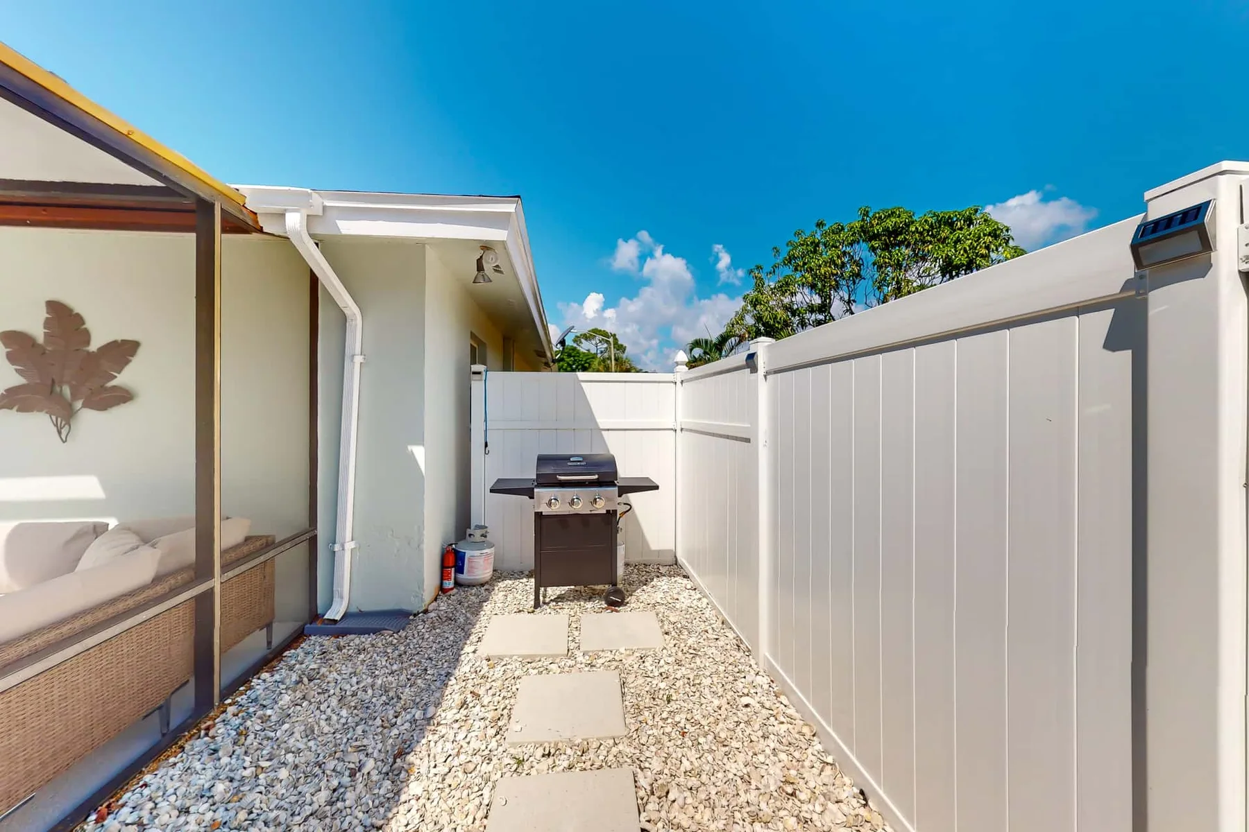 A narrow outdoor patio has gravel, stepping stones, and a tall white fence.