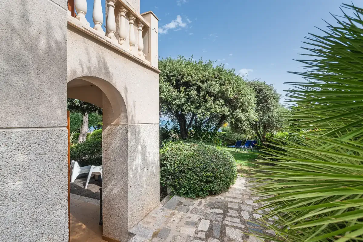 A garden path curves past greenery beside a stucco building with an arched opening.