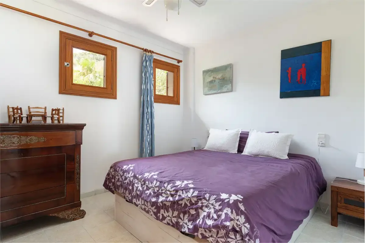 A bedroom with a bed covered in a purple bedspread and framed wall art.