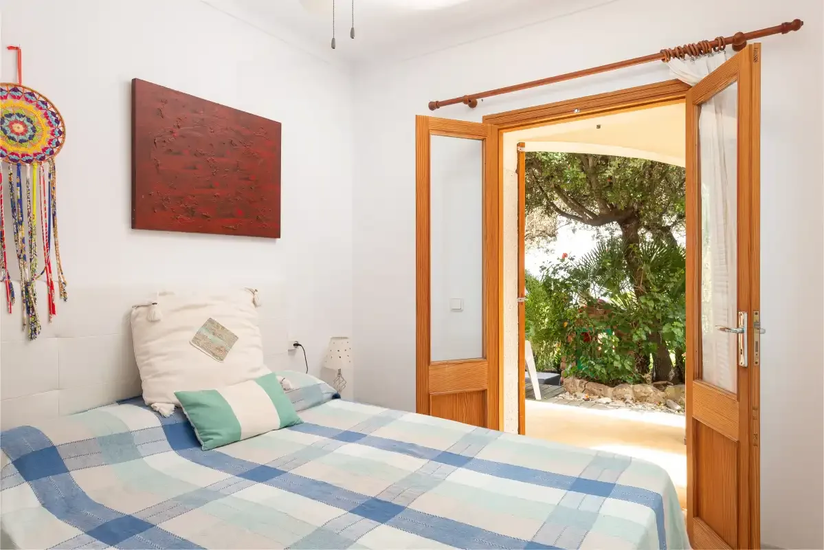 A bedroom with a blue checkered bedspread and open wooden doors to a garden.