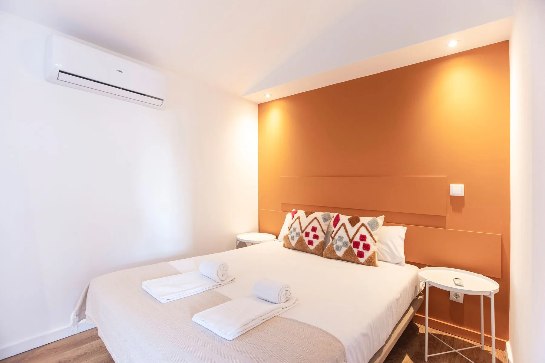 A bedroom with a neatly made bed and patterned cushions against an orange wall.