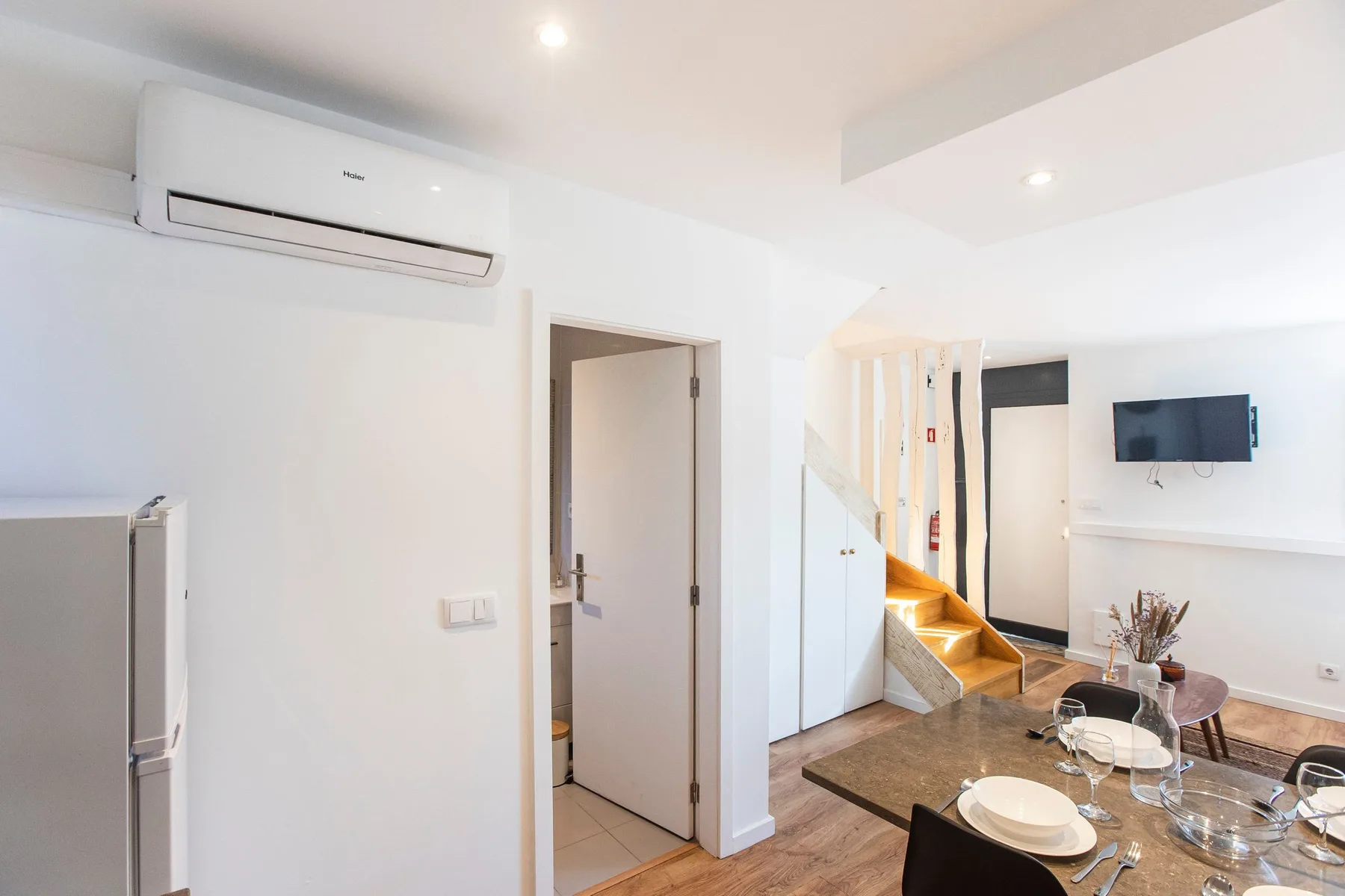 A dining area with a set table sits beside a staircase and wall-mounted television.