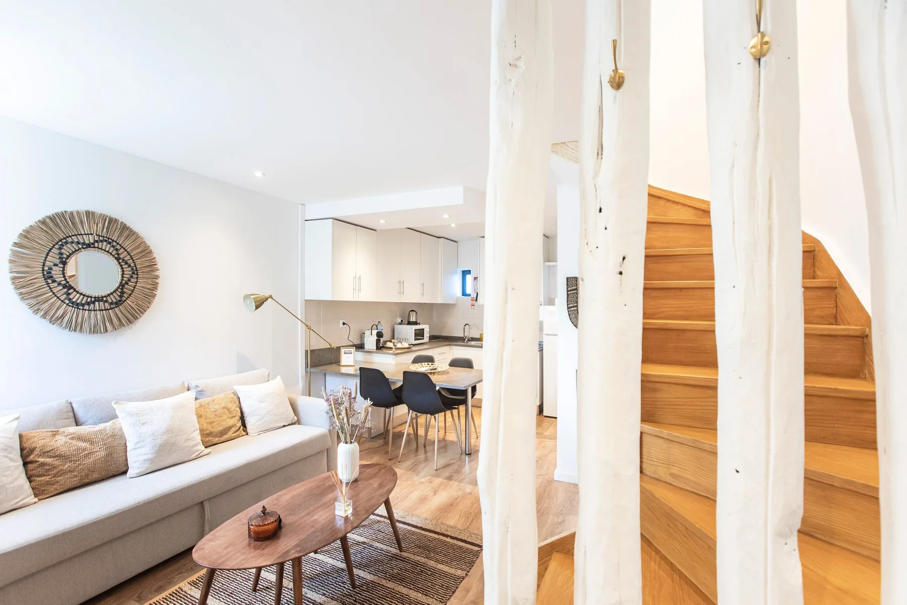 A bright living room opens onto a kitchen, with wooden stairs beside the seating area.