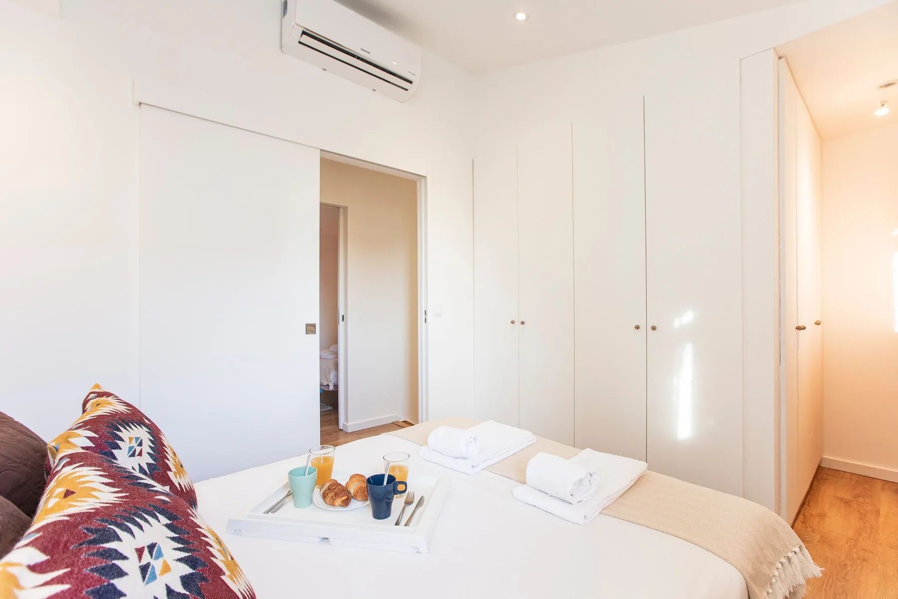 A bright bedroom with a bed, patterned cushions, and white built-in wardrobes.