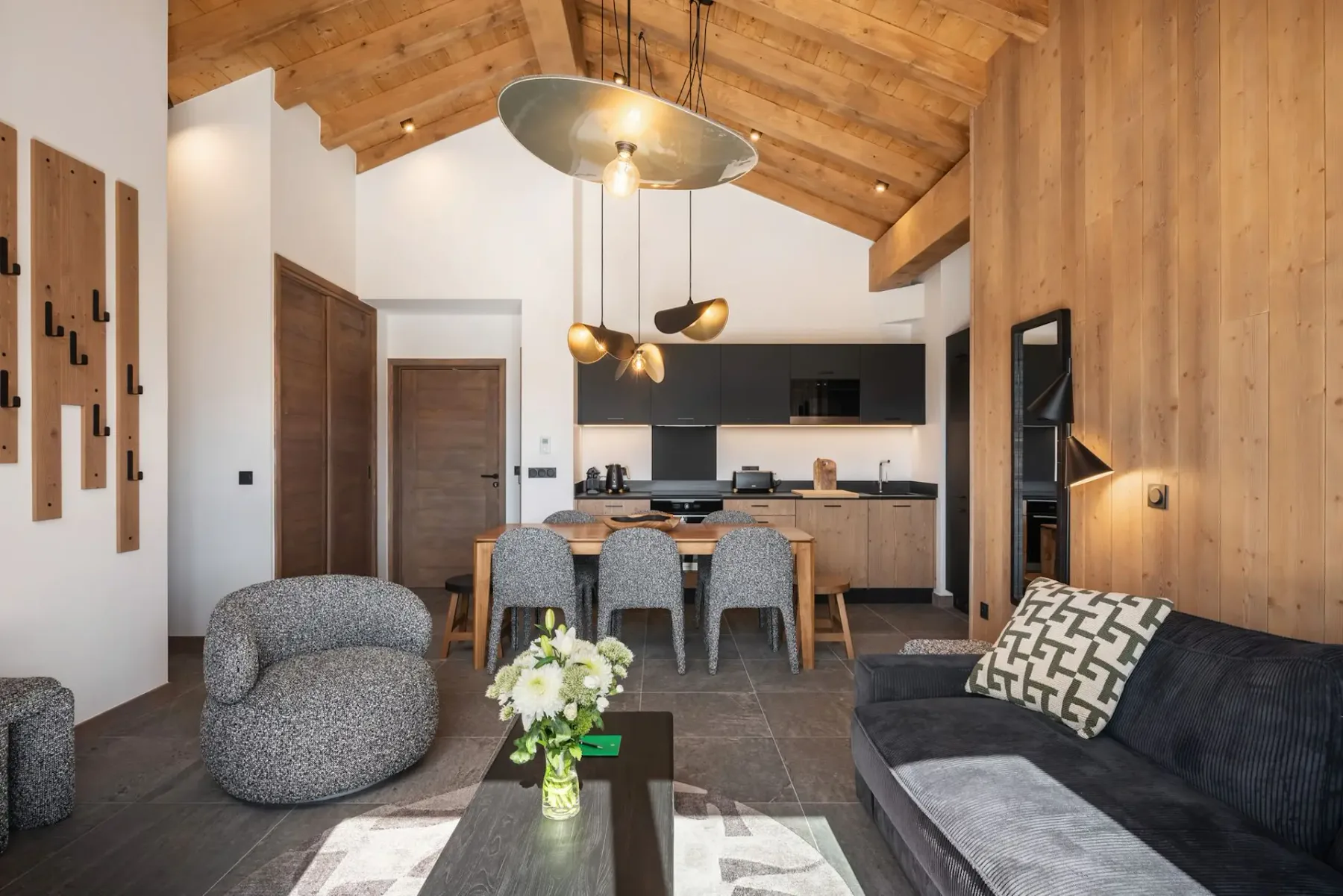 Open-plan living room and kitchen area with wood accents, a dining table, modern appliances, a sofa, and a wall mirror under a beamed ceiling.
