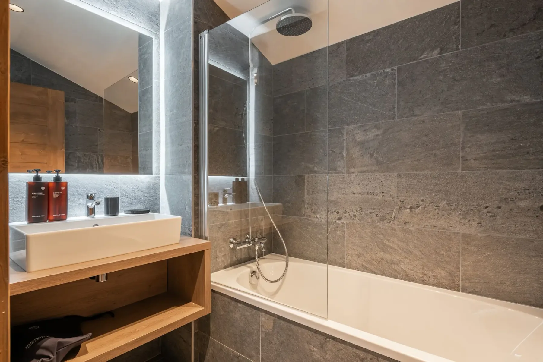 Modern bathroom with gray tiled walls, a bathtub with a glass partition, a white sink, a large mirror, and wooden shelving.