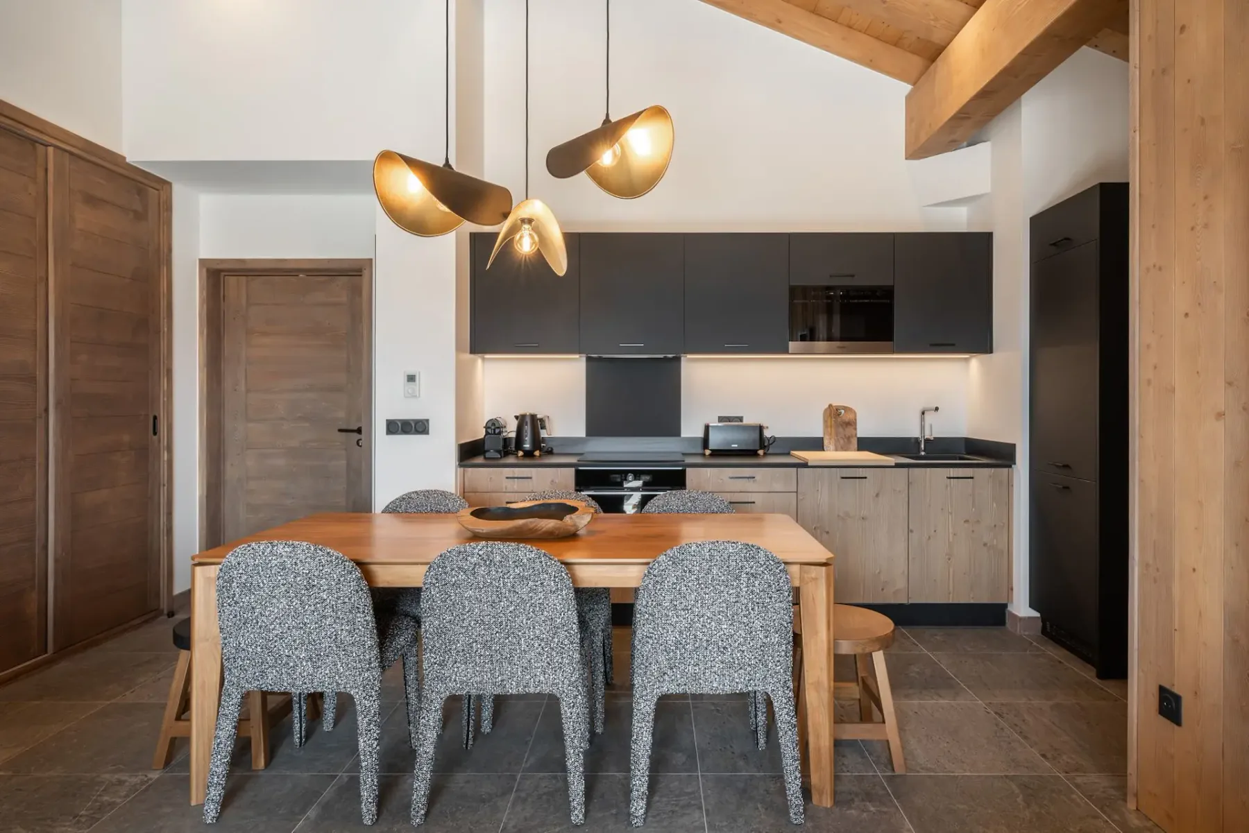 Modern kitchen with wooden ceiling, large wooden dining table, cushioned chairs, gray cabinets, and a unique pendant light.