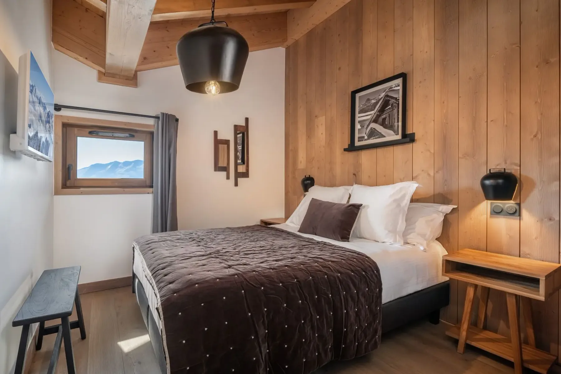 A cozy bedroom with wooden walls, a plush bed, bedside tables, an overhead lamp, and a window view of mountains.