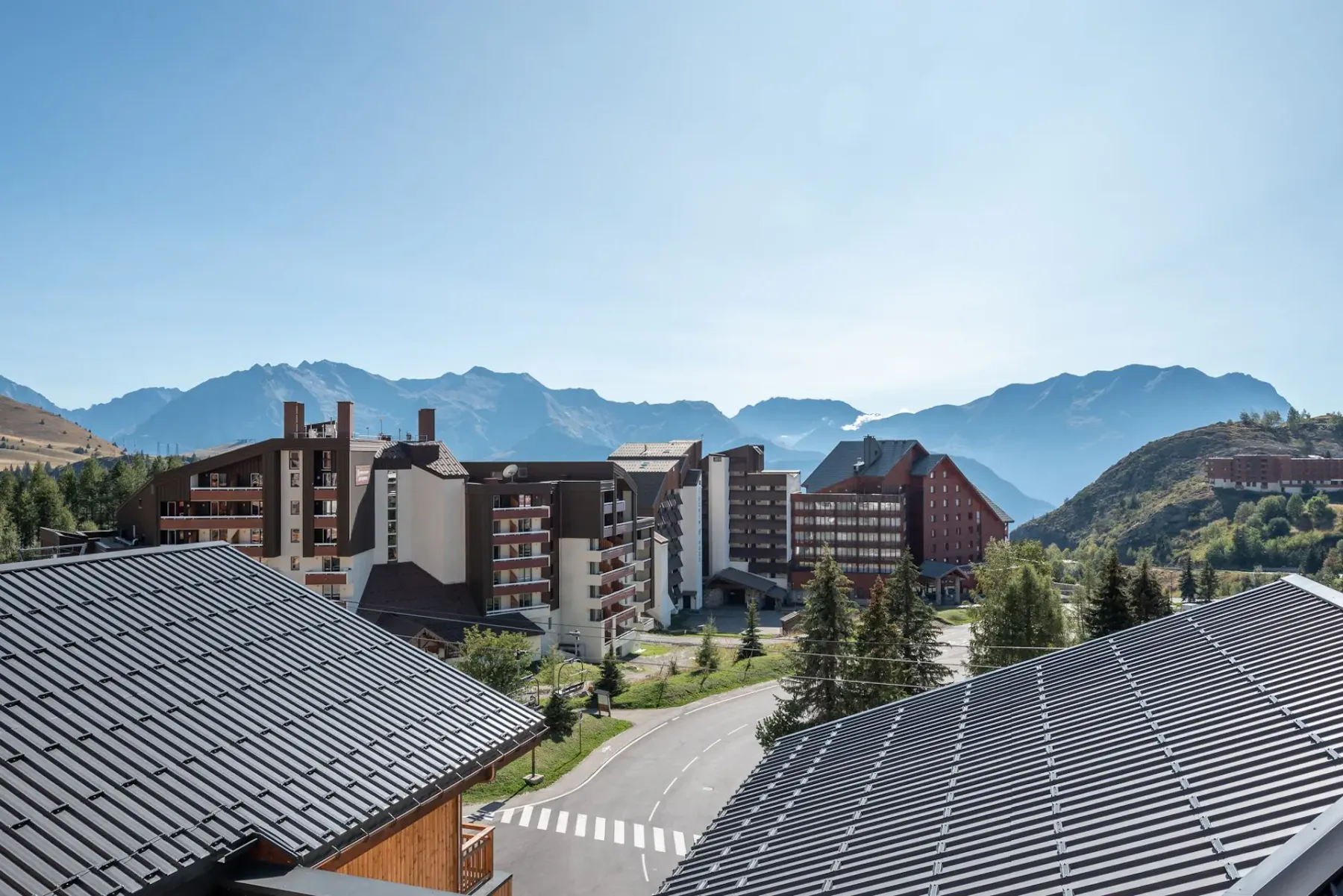 Mountain resort buildings with sloping roofs, surrounded by green trees and set against a backdrop of distant mountain ranges under a clear sky.