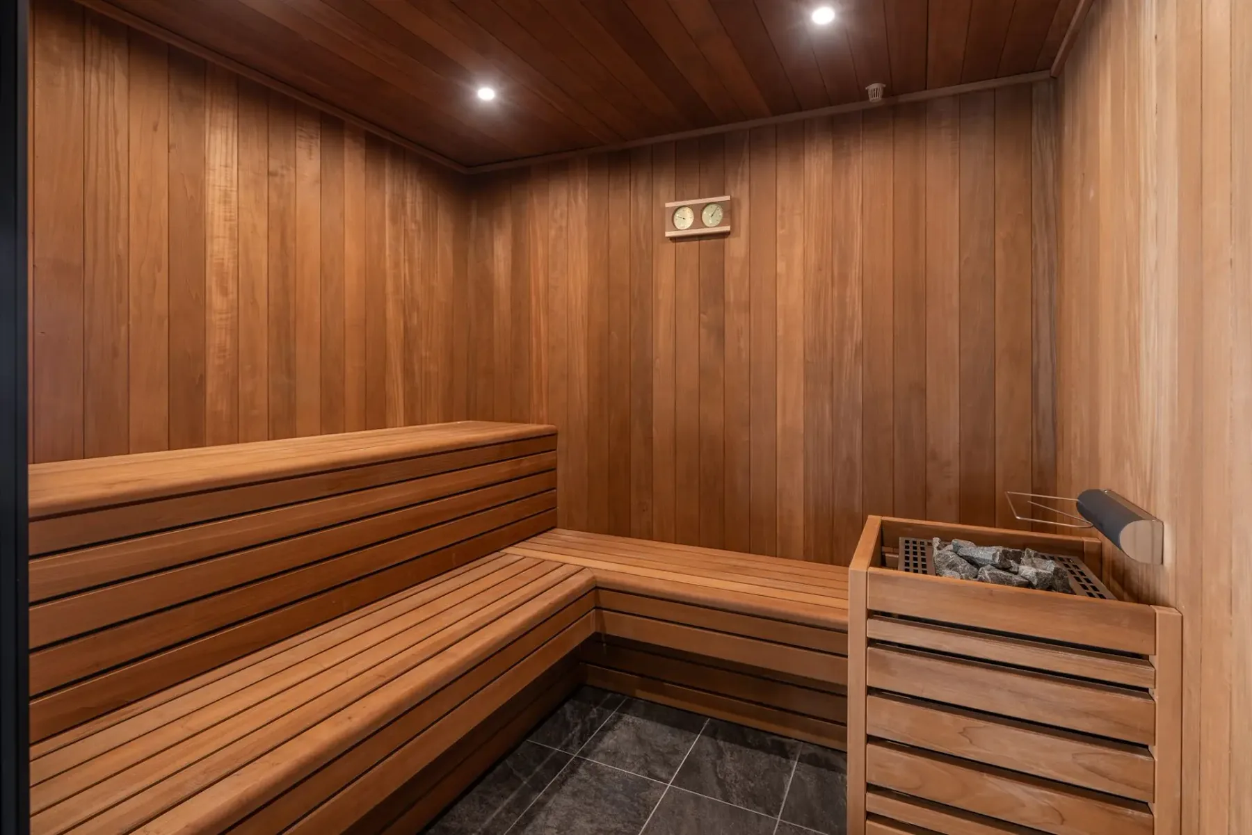 Wooden sauna interior with benches, a small heater with stones, and soft overhead lighting. Wall thermometer and hygrometer present.