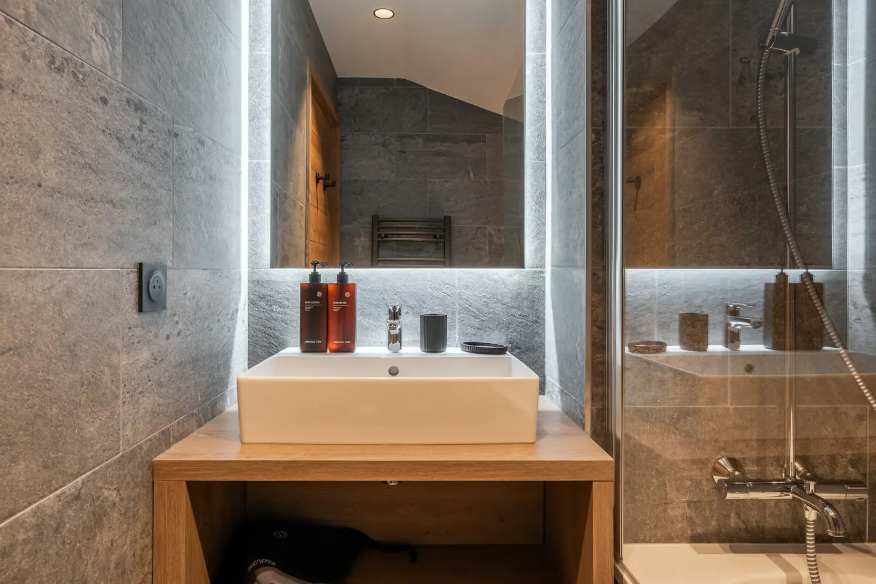 Modern bathroom with gray tiled walls, a bathtub with a glass partition, a white sink, a large mirror, and wooden shelving.