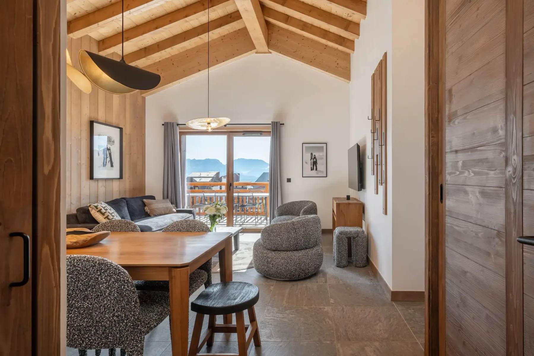 A cozy living room with wooden beams, modern furniture, a dining table, and large windows offering a mountain view.