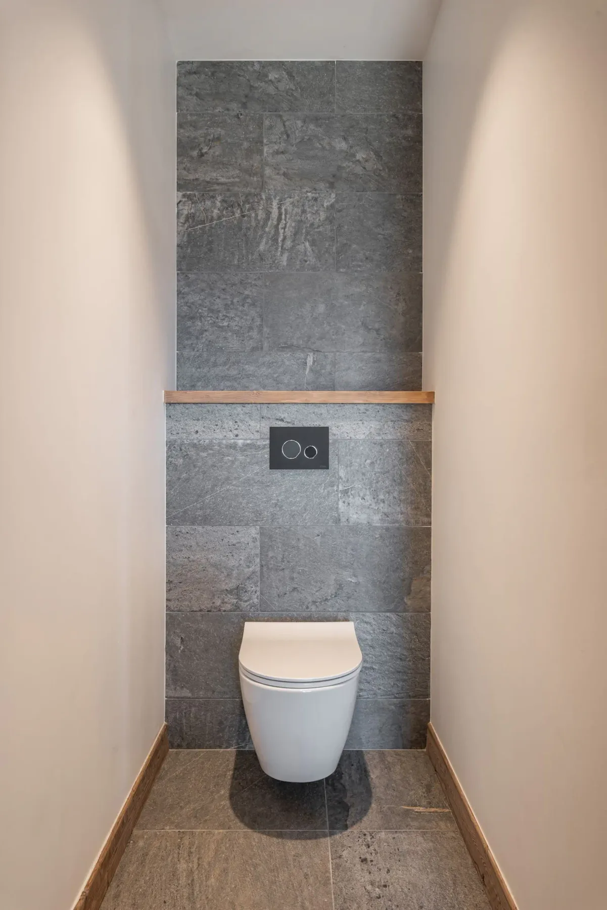 A modern bathroom with a wall-mounted white toilet against a gray tiled accent wall, featuring a dual-flush plate above.