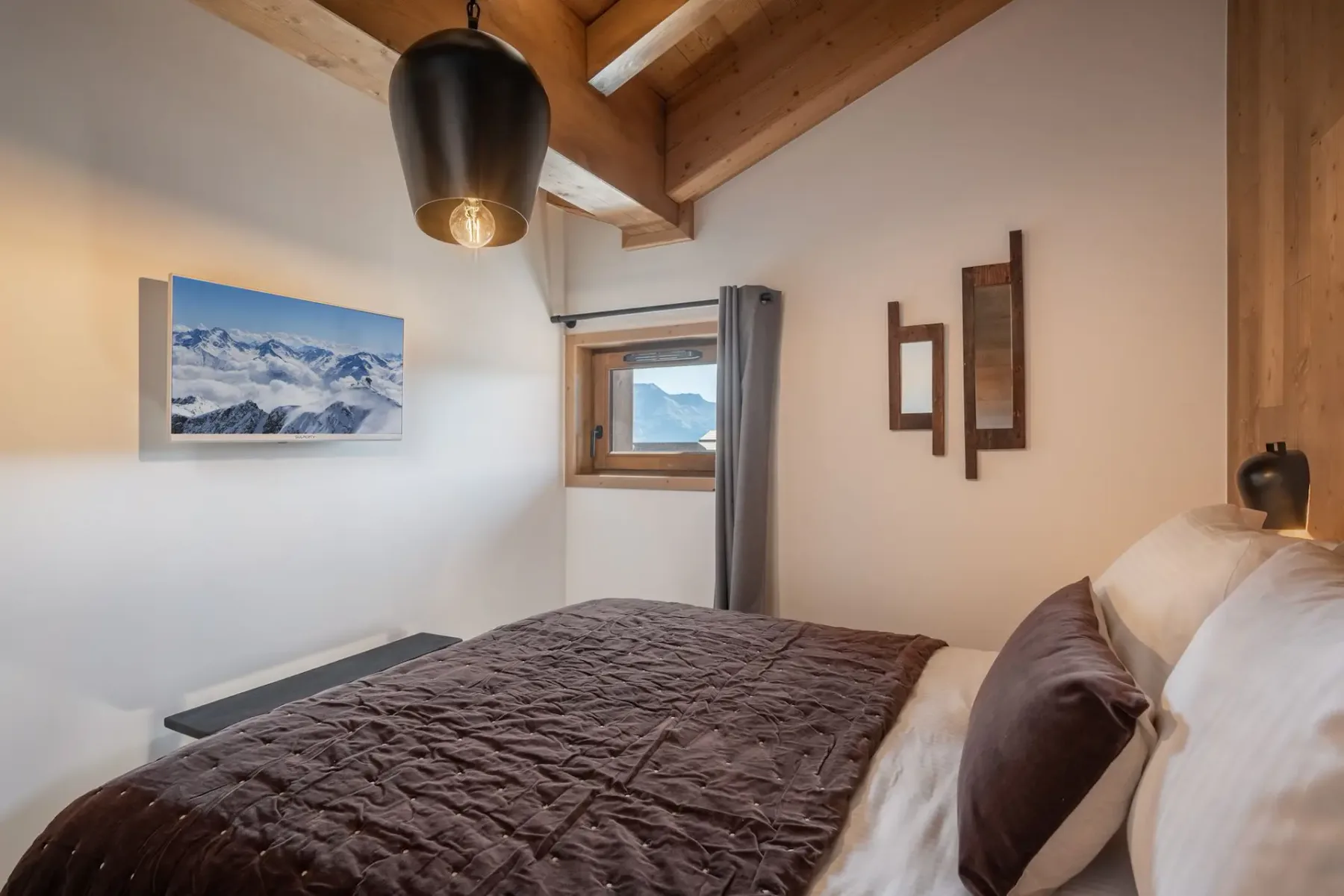 A cozy bedroom with wooden walls, a plush bed, bedside tables, an overhead lamp, and a window view of mountains.