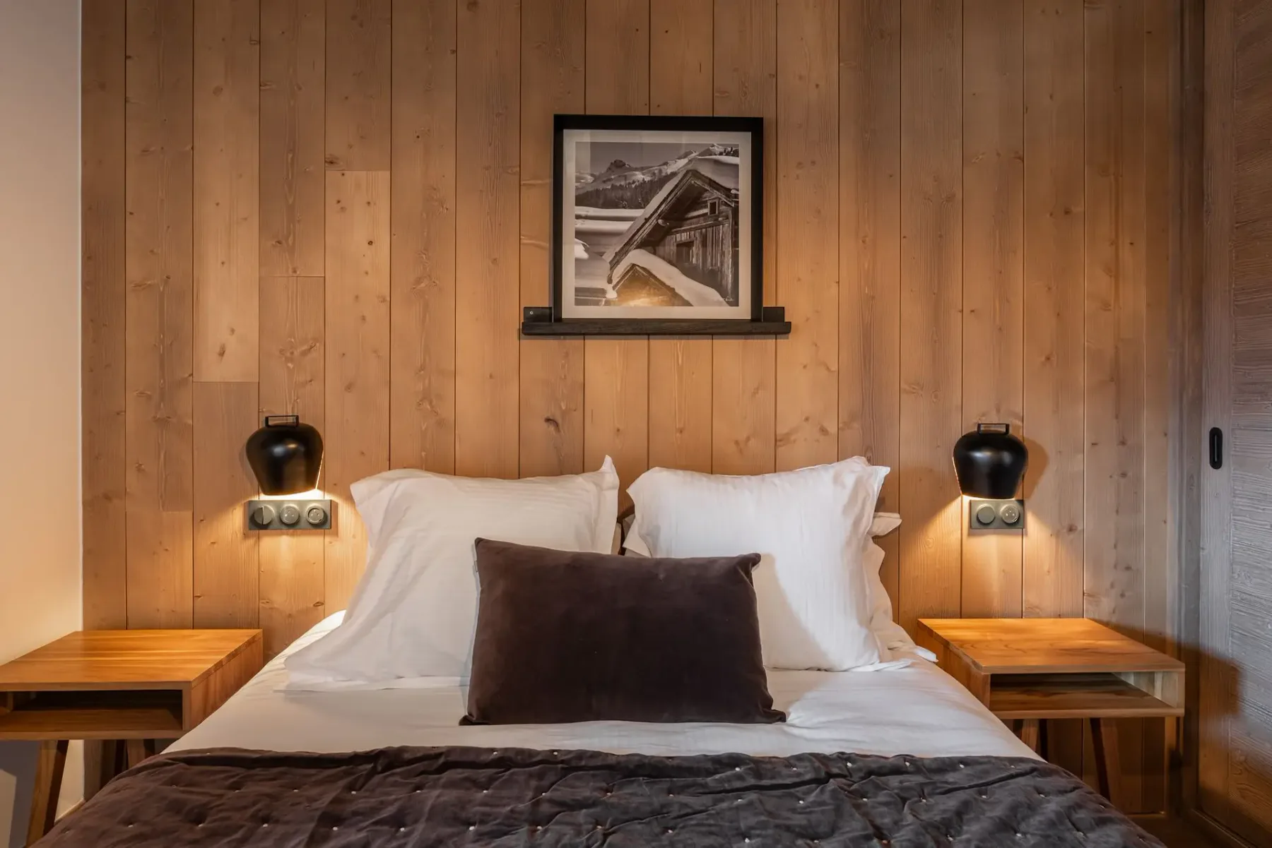 A cozy bedroom with wooden walls, a plush bed, bedside tables, an overhead lamp, and a window view of mountains.