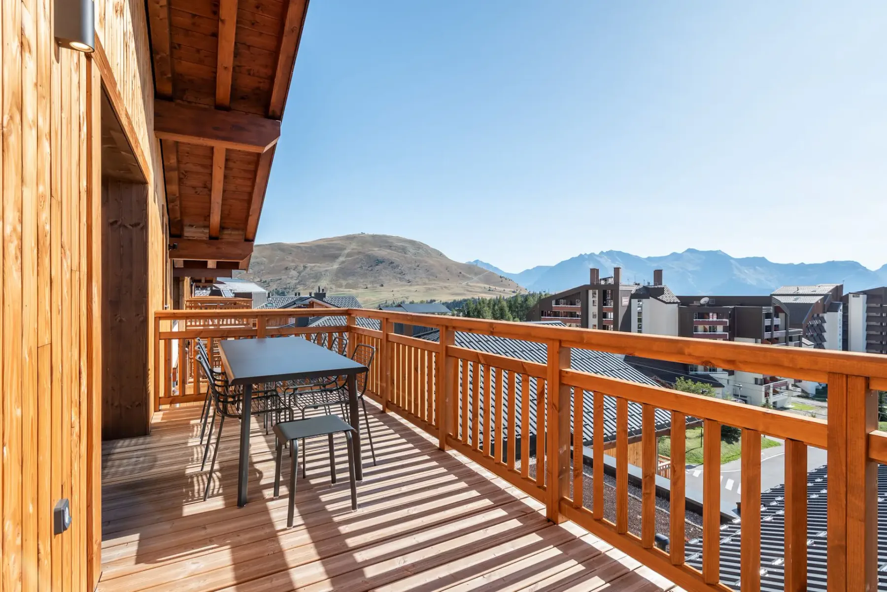 Wooden balcony with a dining table, chairs, and mountain view on a sunny day. Nearby buildings and clear blue sky in the background.