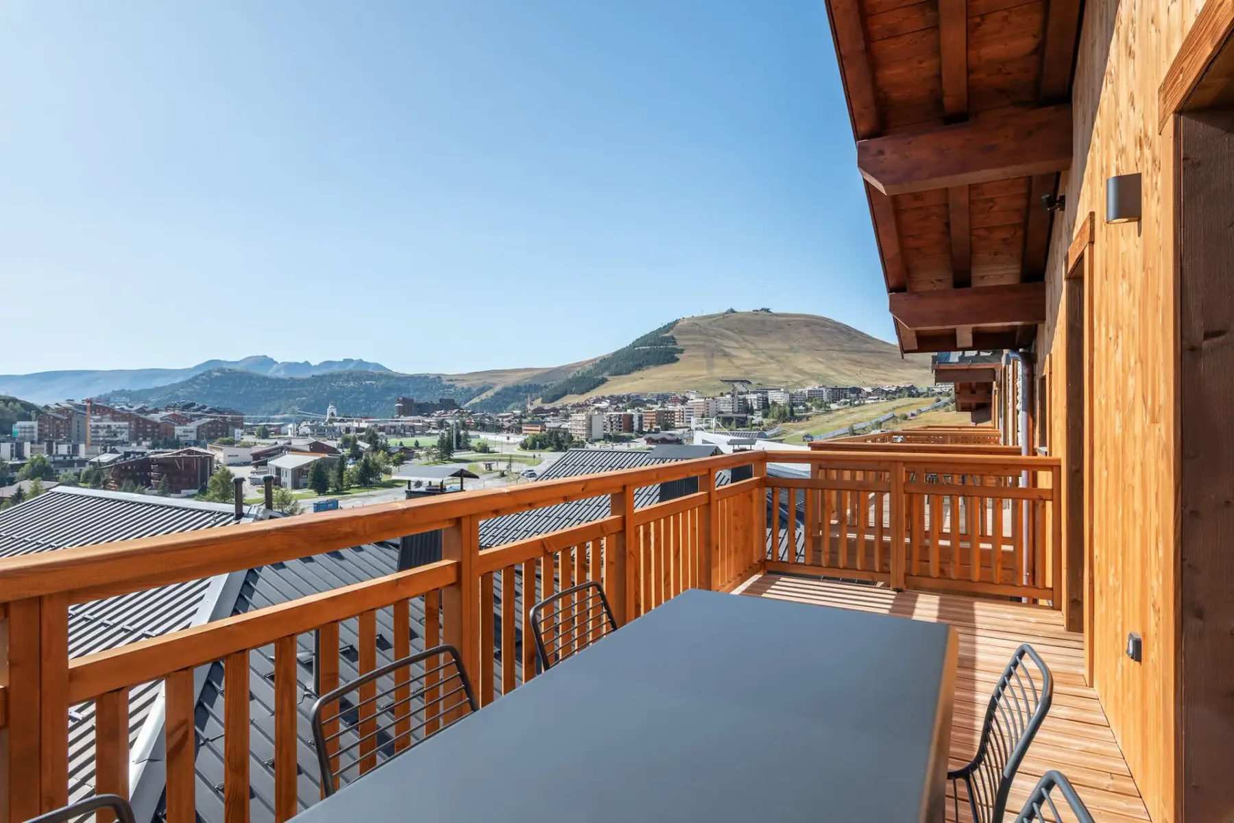 Wooden balcony with a dining table, chairs, and mountain view on a sunny day. Nearby buildings and clear blue sky in the background.