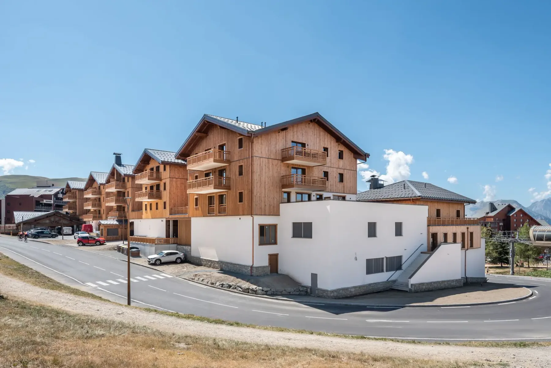 Wooden alpine-style apartment buildings with balconies under a clear blue sky, cars parked in front, on a quiet road with a mountain backdrop.
