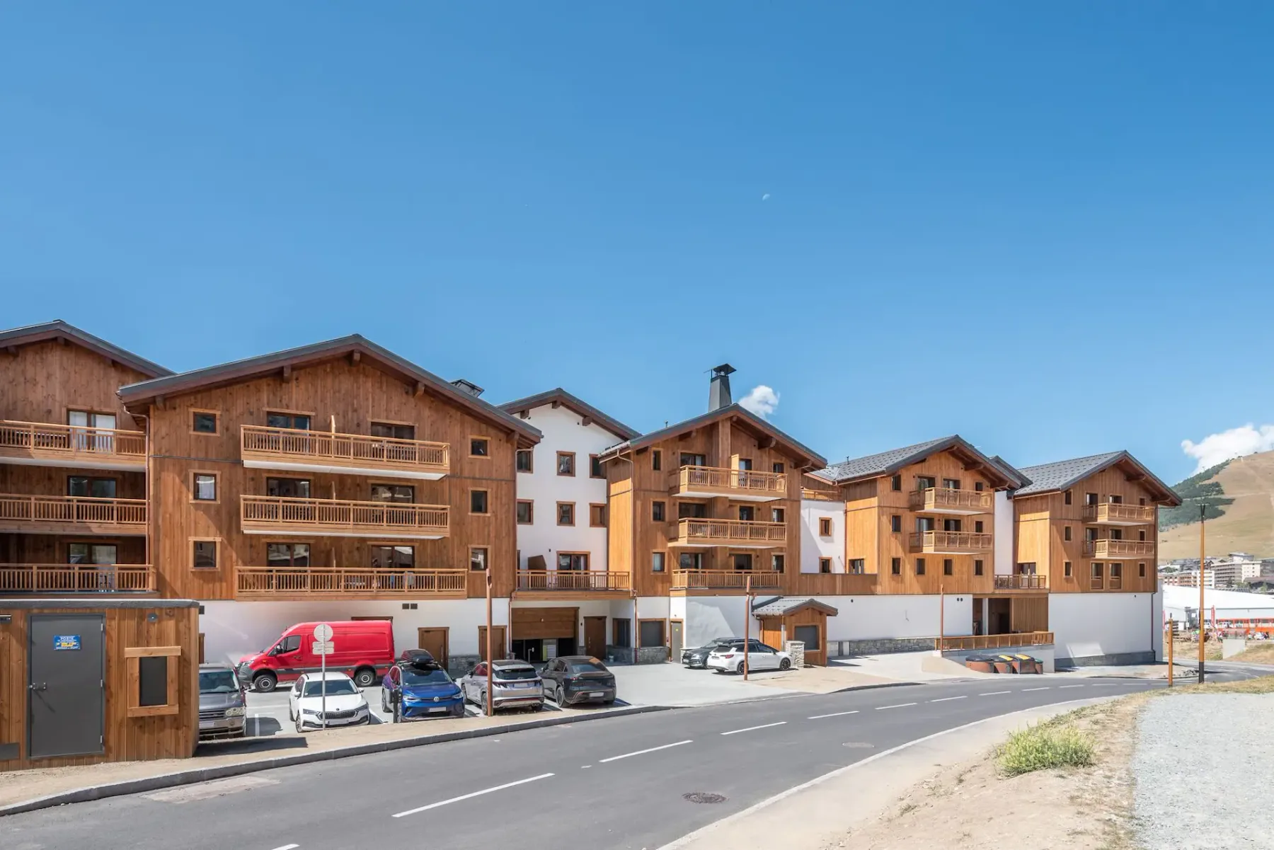 Wooden alpine-style apartment buildings with balconies under a clear blue sky, cars parked in front, on a quiet road with a mountain backdrop.