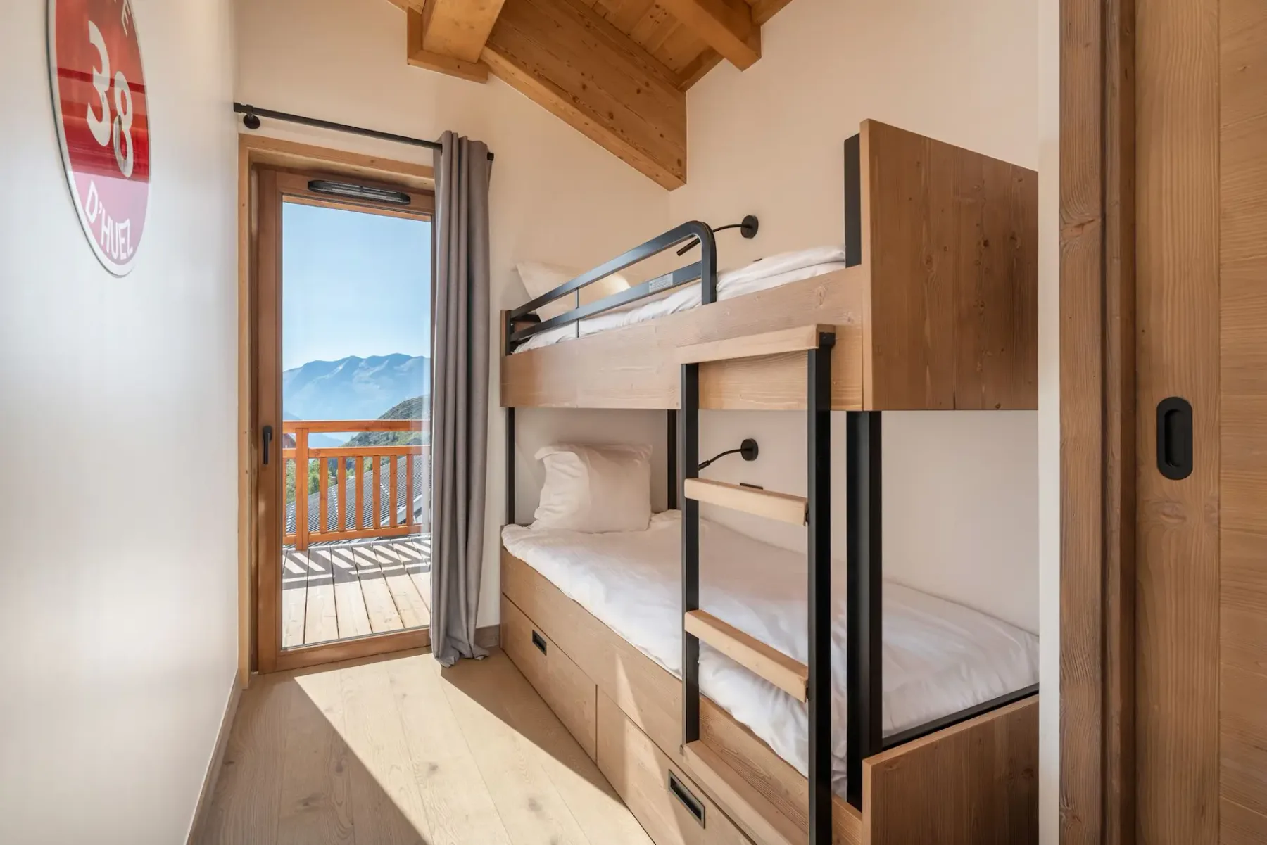 Narrow room with wooden bunk beds, wooden ceiling, and a door leading to a balcony with mountain views. Daylight fills the space.