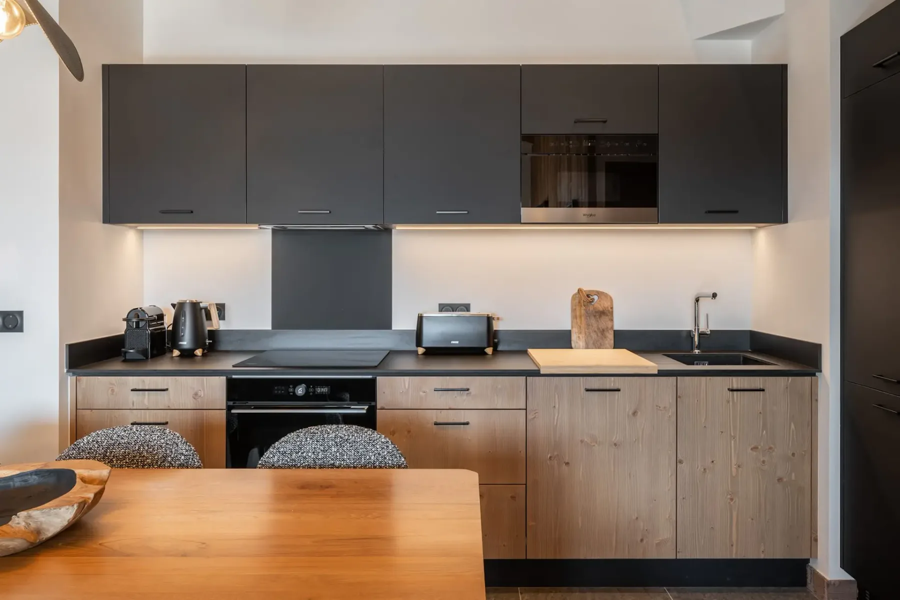 Modern kitchen with wooden ceiling, large wooden dining table, cushioned chairs, gray cabinets, and a unique pendant light.