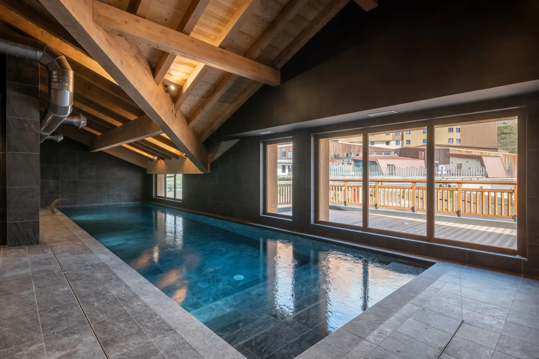 Indoor swimming pool under a vaulted wooden ceiling with natural light streaming through large windows showing a view of an outdoor deck.