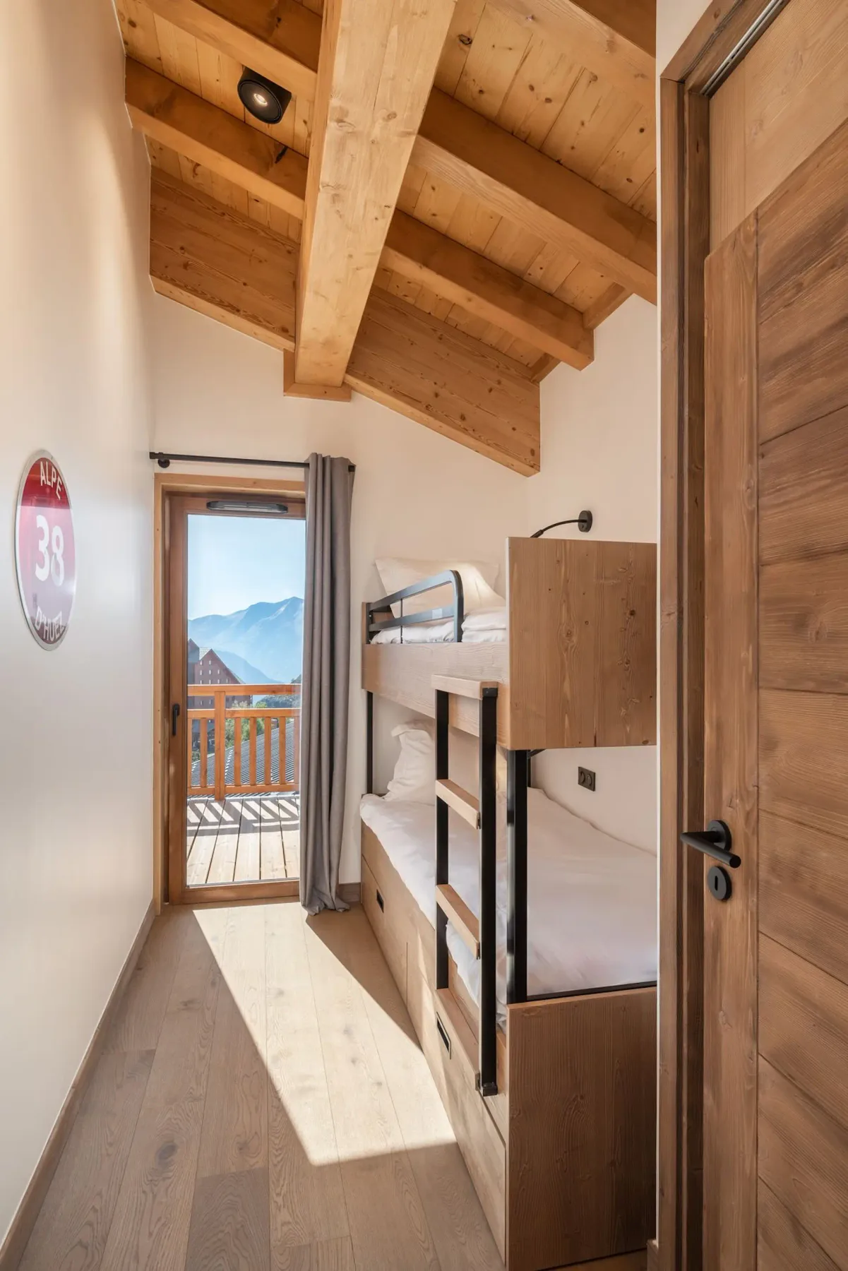 Narrow room with wooden bunk beds, wooden ceiling, and a door leading to a balcony with mountain views. Daylight fills the space.
