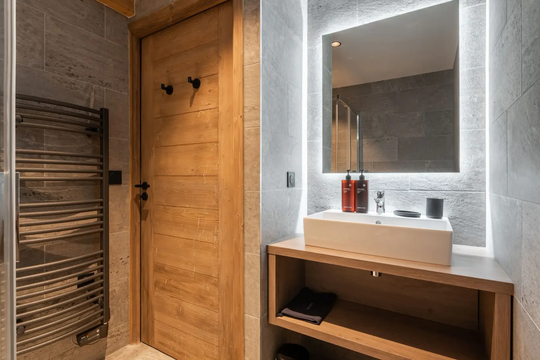 Modern bathroom with a wooden door, illuminated mirror, white rectangular sink, toiletries, heated towel rack, and stone tile walls.