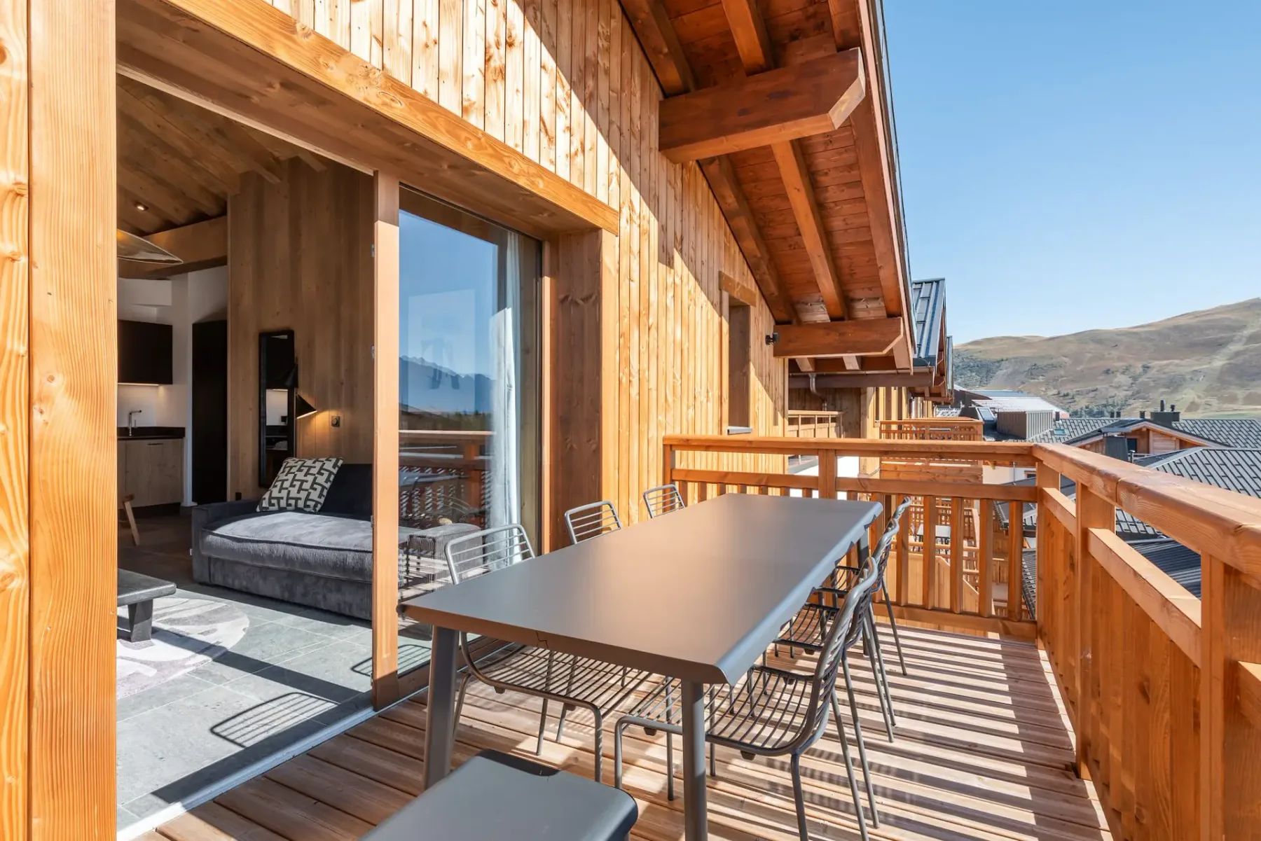 Wooden balcony with a dining table, chairs, and mountain view on a sunny day. Nearby buildings and clear blue sky in the background.
