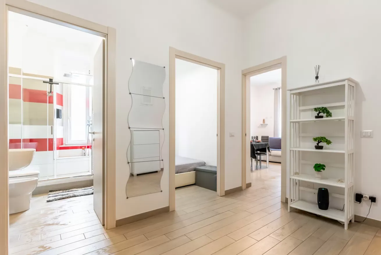 Un pasillo interior luminoso da a un baño y habitaciones contiguas, con una estantería blanca.