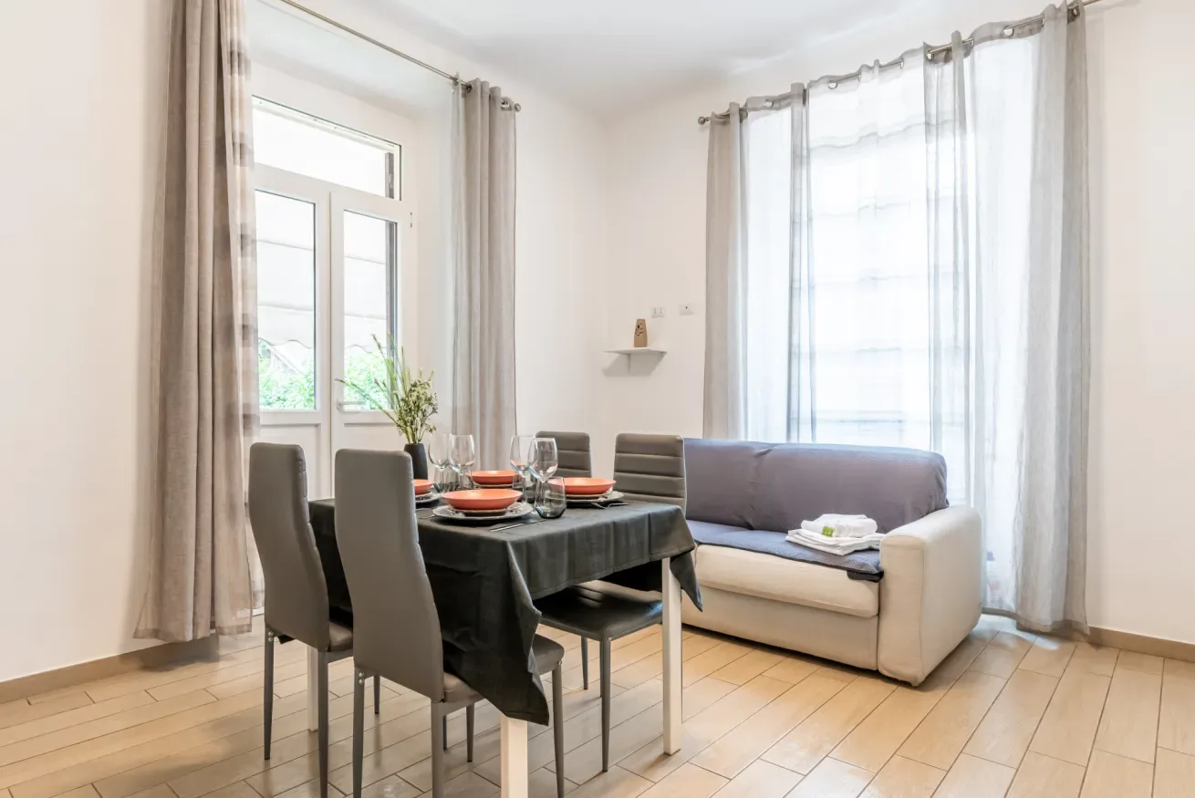 Una luminosa zona de comedor y estar, con una mesa junto a un sofá y grandes ventanas.