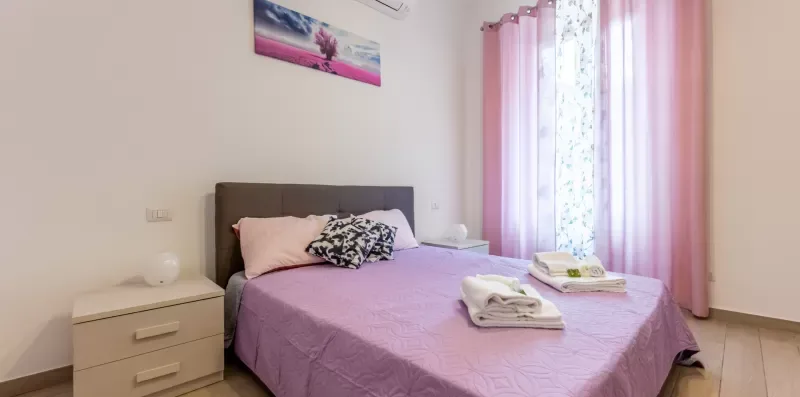 Un dormitorio con una cama vestida con ropa de cama lila junto a una ventana con cortinas rosas.