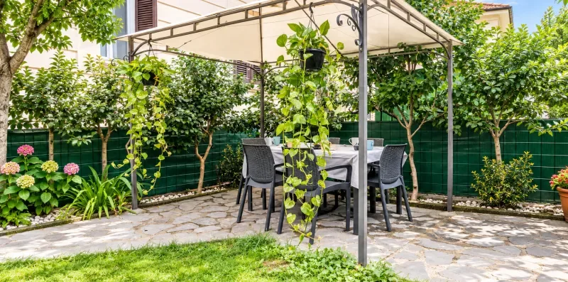 Un patio de jardín con asientos al aire libre bajo un toldo, rodeado de vegetación.