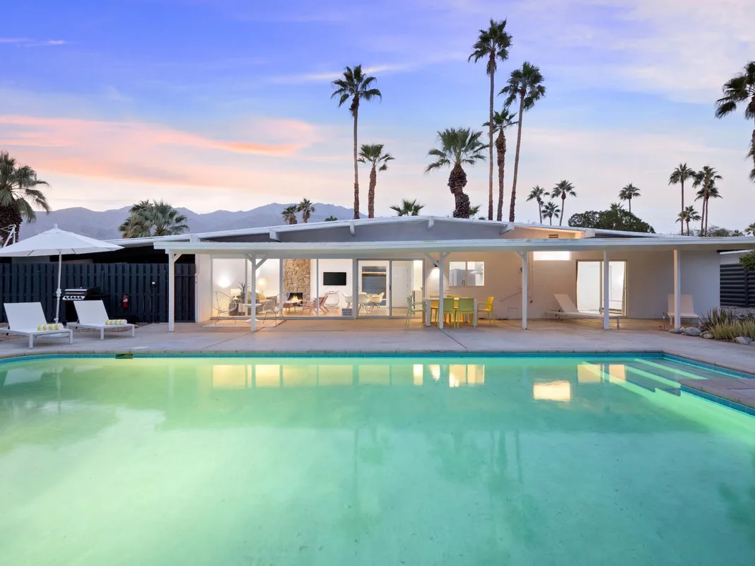 Una piscina frente a una casa moderna con puertas de vidrio y palmeras.