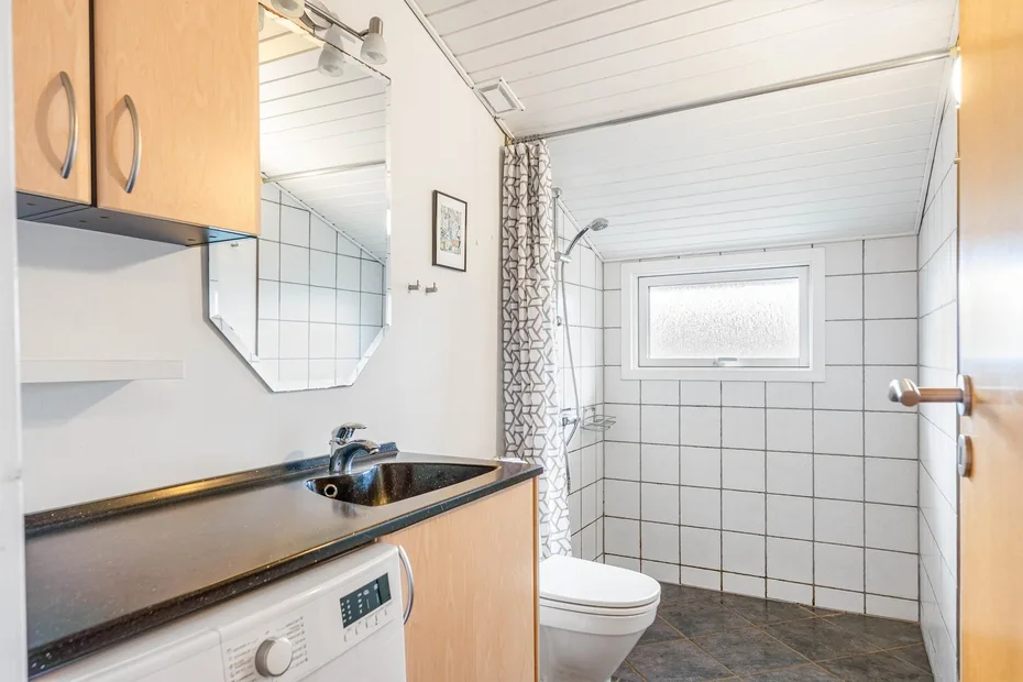 Un baño alicatado con lavabo, inodoro y zona de ducha bajo una ventana pequeña.