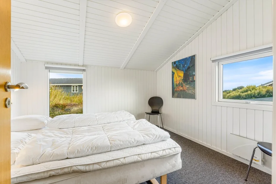 Un dormitorio luminoso con una cama baja, ropa de cama blanca y ventanas.