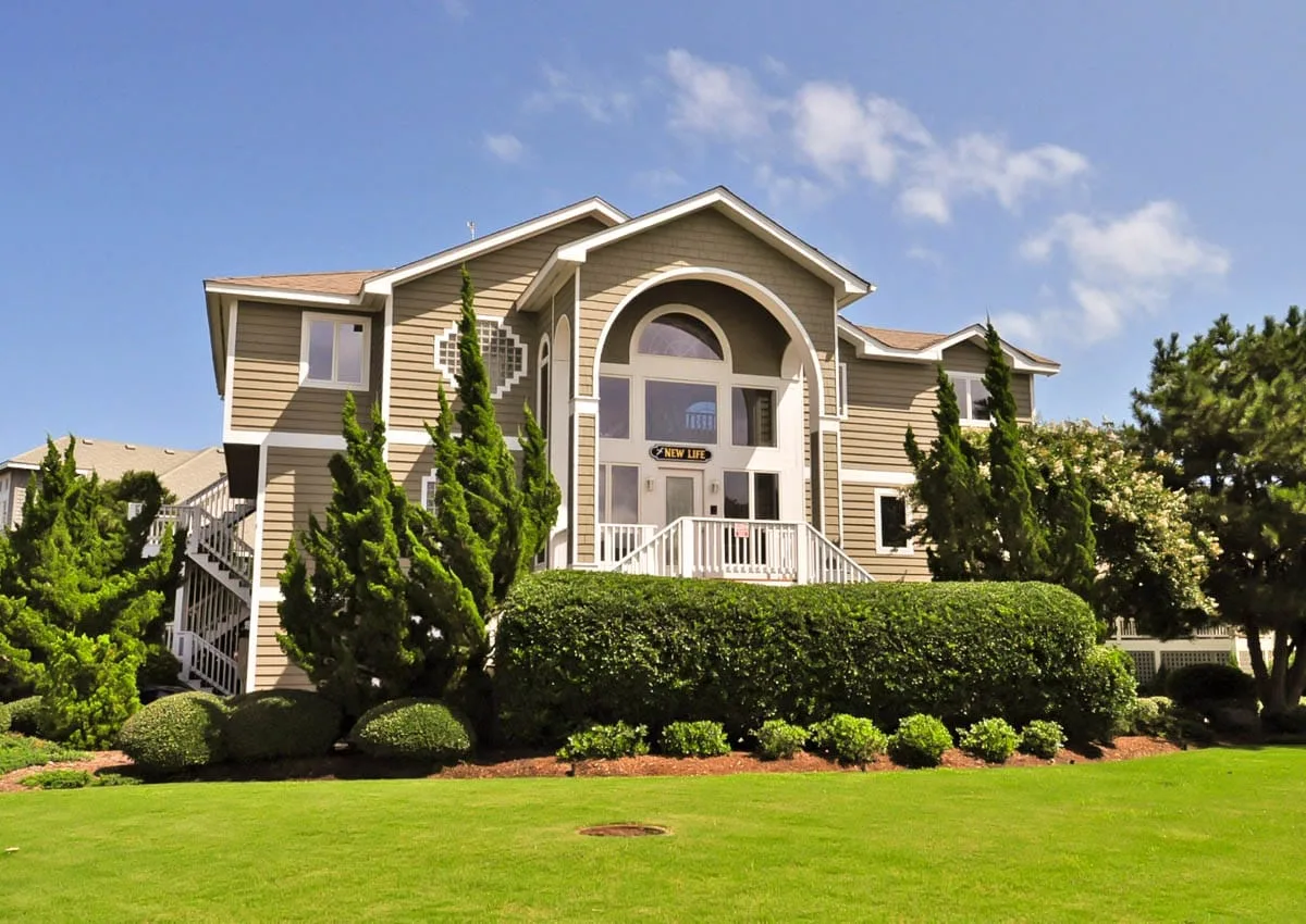 A large house with a covered entrance stands behind a trimmed hedge and lawn.