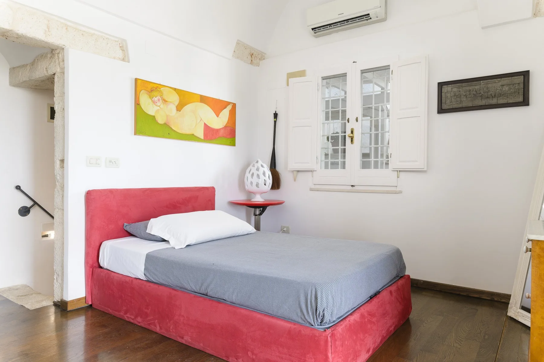 Un dormitorio con una cama roja bajo una ventana con postigos abiertos.