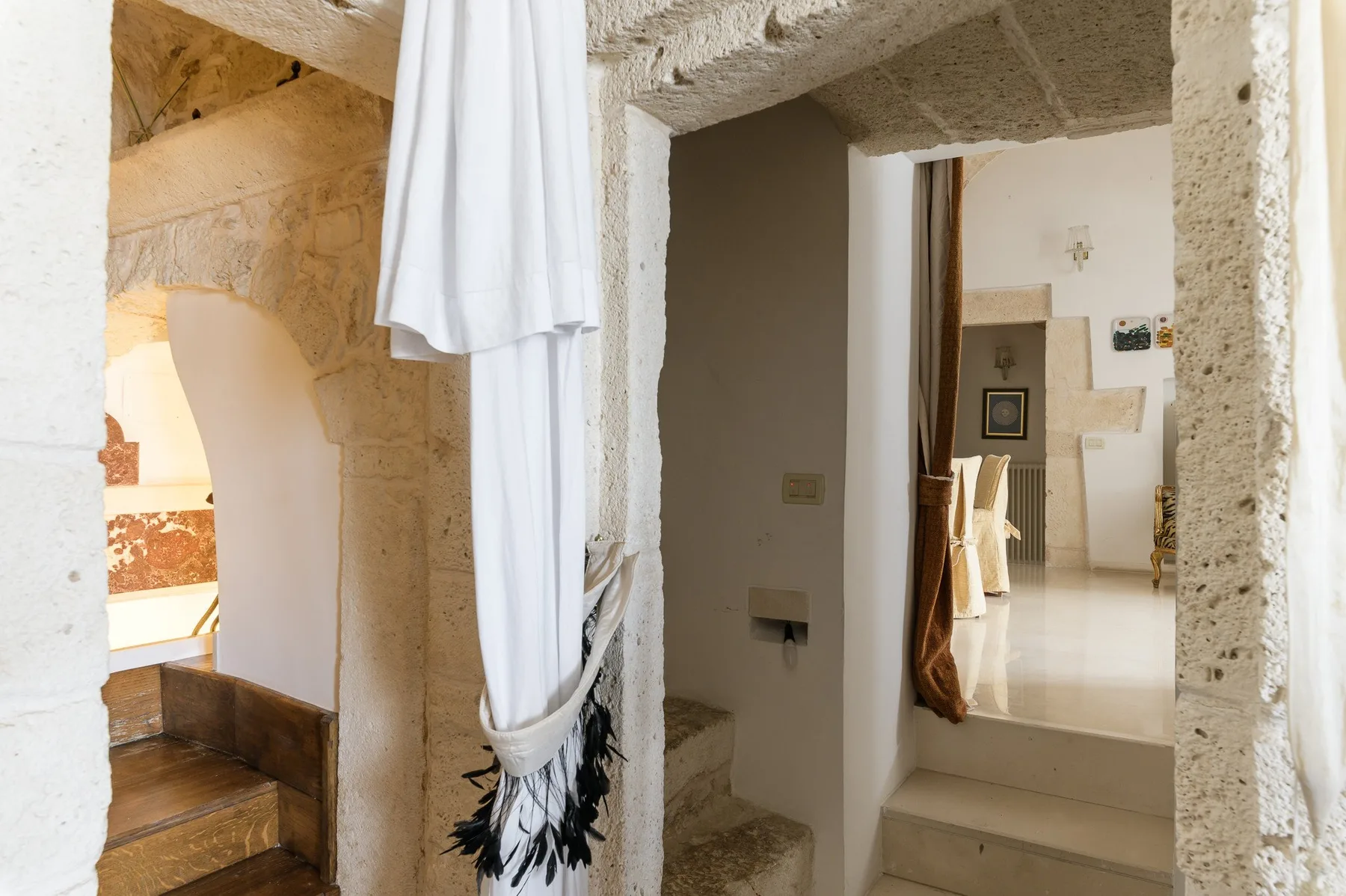 Un pasillo interior de piedra se abre a una habitación más luminosa, con una cortina blanca junto a los escalones.