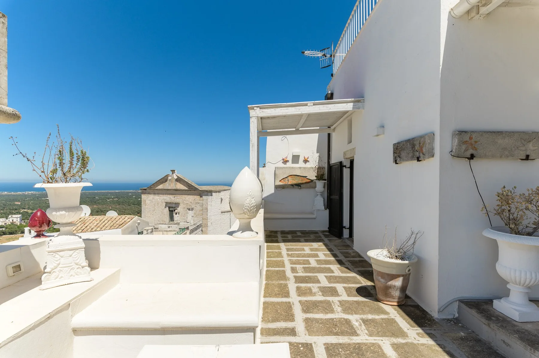 Una terraza soleada con asientos blancos, plantas en macetas y vistas al mar.
