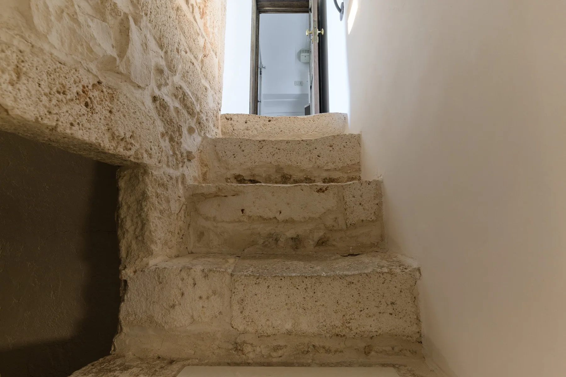 Una estrecha escalera de piedra sube hasta una puerta de vidrio junto a una pared clara.