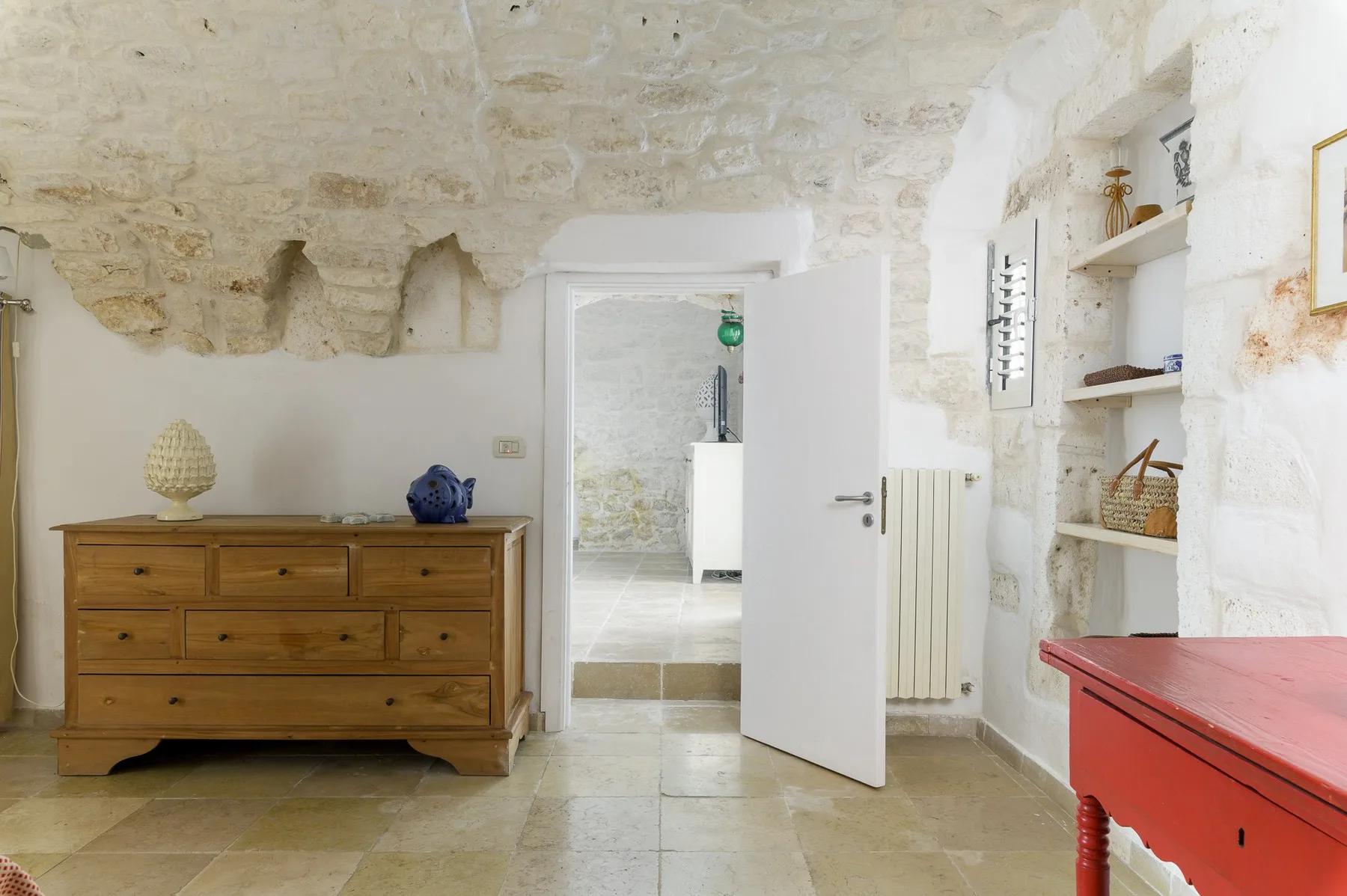 Una habitación con paredes de piedra, una cómoda de madera, una puerta abierta y una mesa roja.