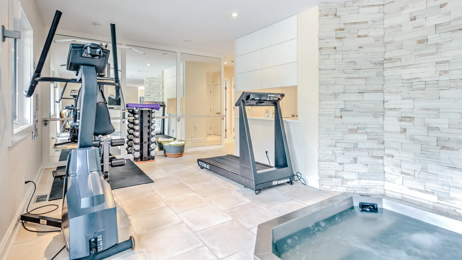 A bright indoor gym with exercise equipment beside a small tiled pool.