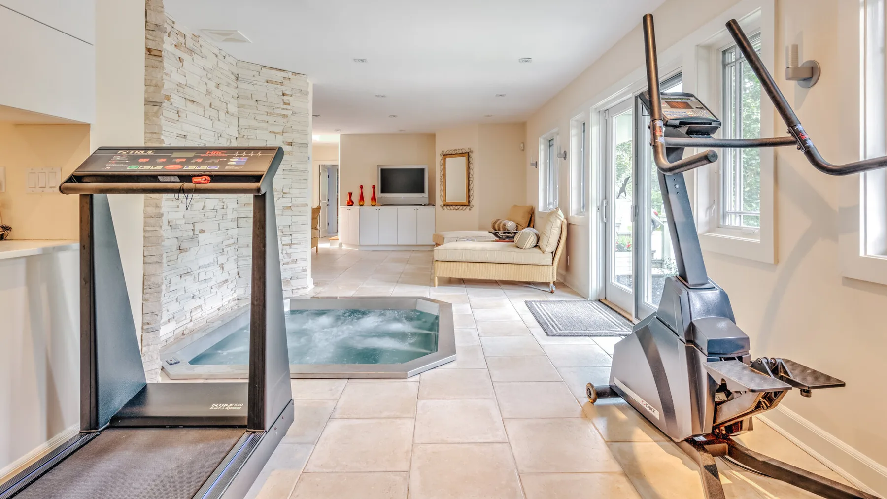 A bright indoor exercise and relaxation room with fitness equipment and a sunken spa.