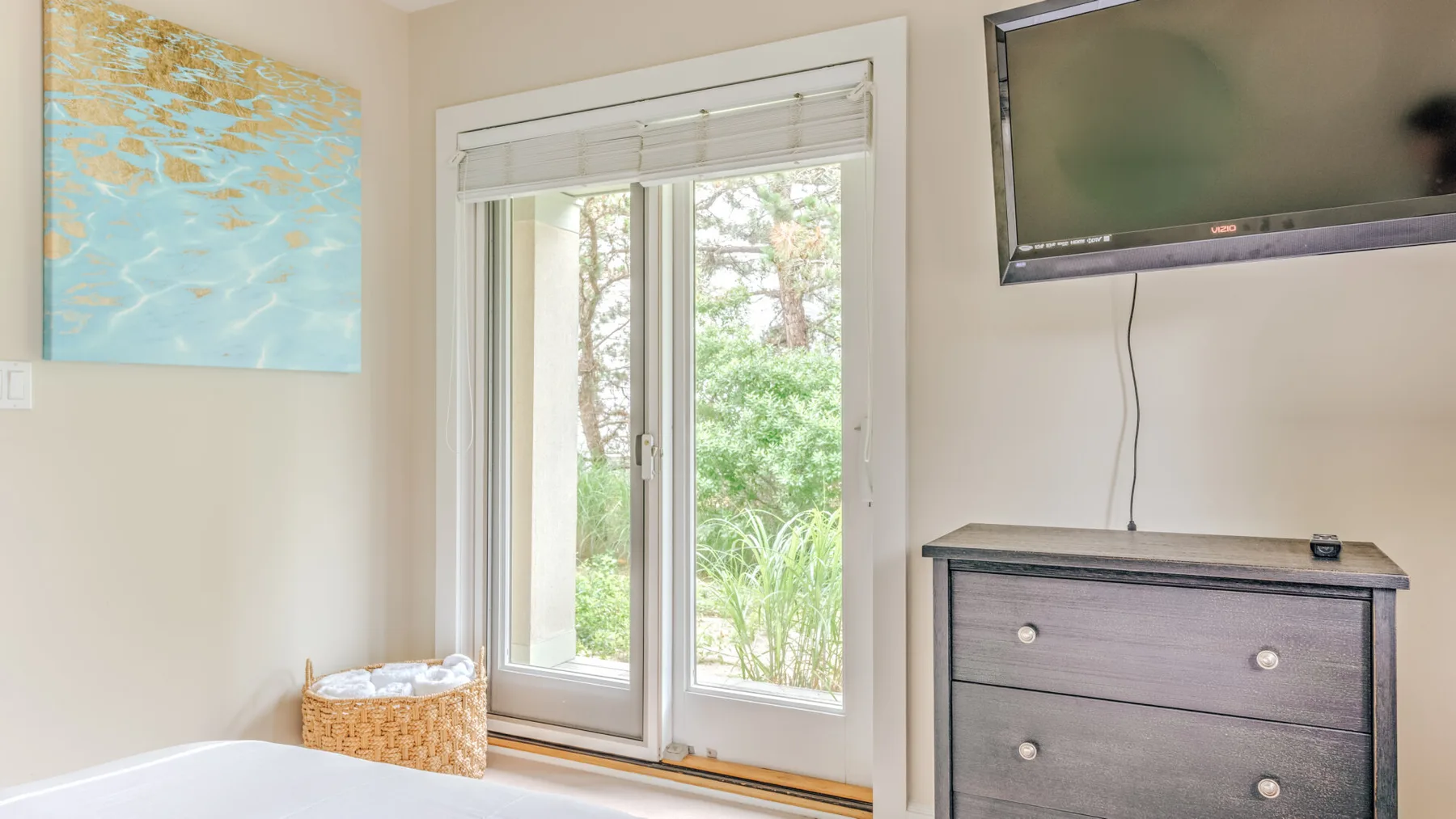 A bedroom corner with a wall-mounted television, dresser, and glass doors overlooking greenery.